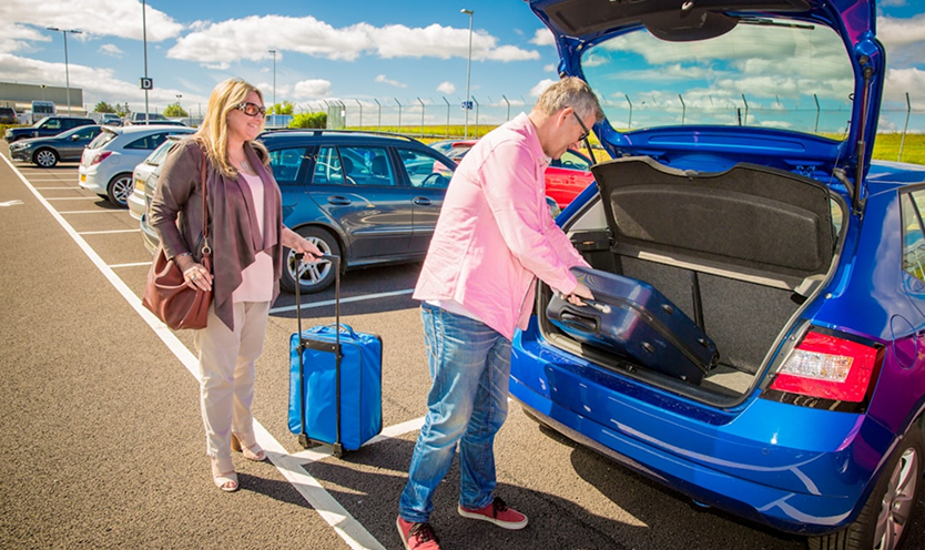A man in a pink shirt loads a large blue suitcase into the trunk of a blue hatchback while a woman with a rolling suitcase stands nearby in an outdoor airport parking lot.