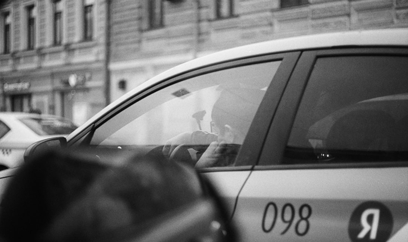 A candid black-and-white close-up of a taxi or ride-service vehicle in a city, with the driver visible through the window and service numbers on the car door.